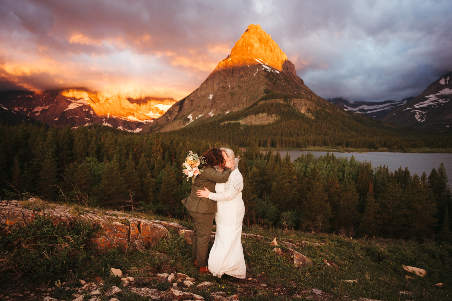A couple embracing during sunrise on their elopement day in Glacier National Park. Behind them is a mountain lit up orange by the morning sun.