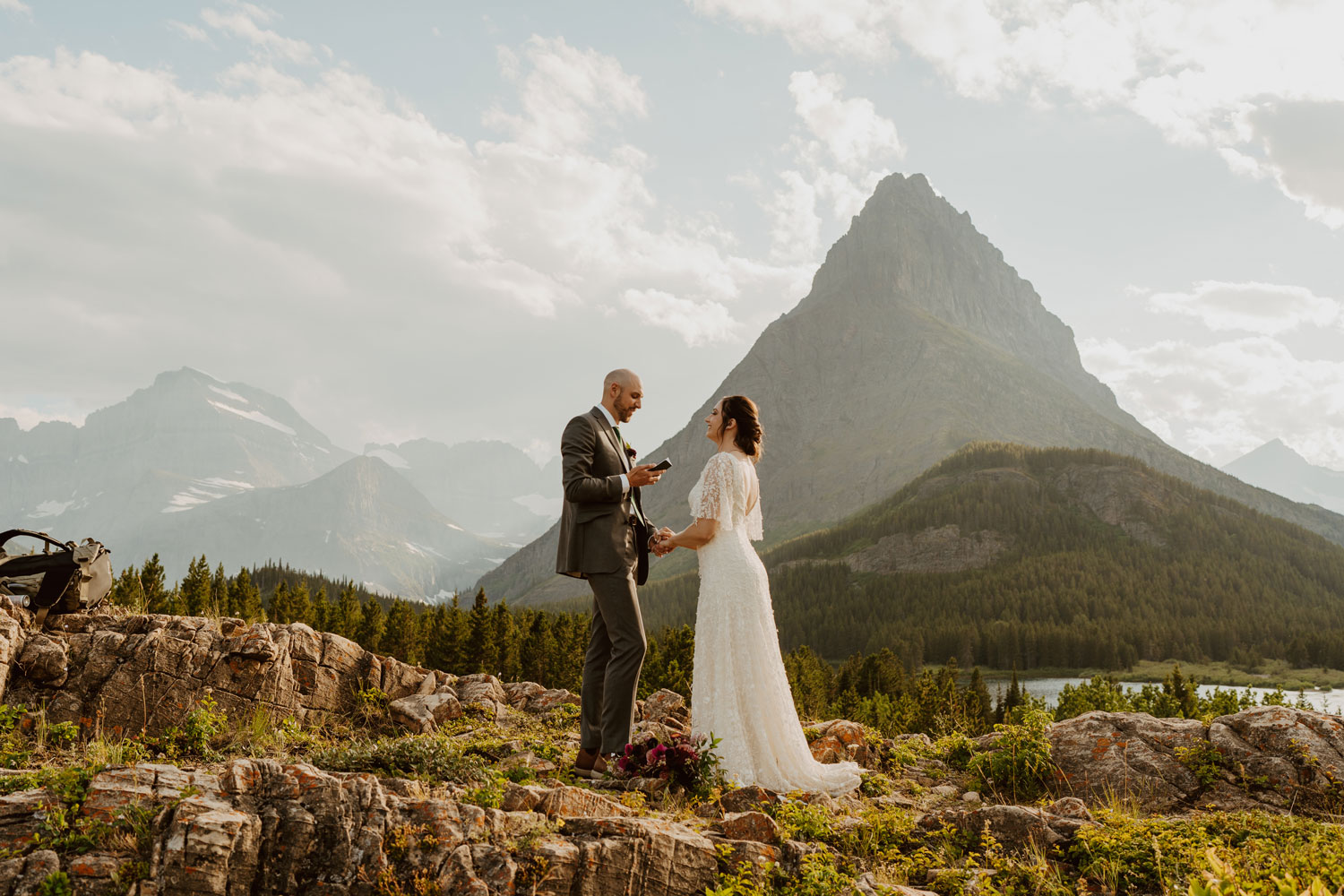 Couple exchanging vows during their Glacier National Park elopement standing on rocky terrain with a mountainscape behind them and a cloudy sky.